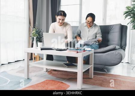 Zwei Frauen arbeiten zu Hause mit einem Laptop und gedruckten Dokumenten, prüfen Papiere, Notizen und Haushaltspläne in einem hellen Wohnzimmer und zeigen sie Stockfoto