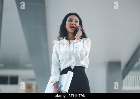 Tiefwinkelansicht einer professionellen Geschäftsfrau, die mit einem Smartphone spricht, während sie Berichte und einen Stift in einem modernen Bürogebäude hält, die den Kunden repräsentiert Stockfoto