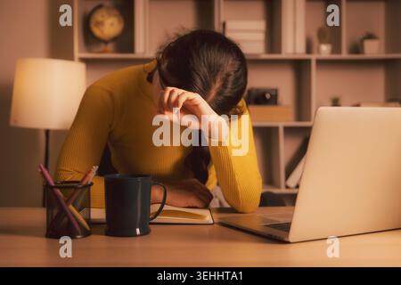 Erschöpfte junge asiatische Frau, die die Augen bedeckt, während sie nachts am Laptop im gemütlichen Heimbüro sitzt, Burnout illustriert, Stress studiert, Müdigkeit am Arbeitsplatz, tot Stockfoto