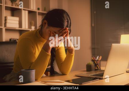 Erschöpfte junge asiatische Frau, die das Gesicht bedeckt, während sie nachts am Laptop im gemütlichen Home Office sitzt, Burnout, Angst, Fernarbeit Stress, Tote ausdrückt Stockfoto