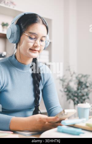 Junge asiatische Frau in Brille mit Smartphone beim Lernen mit Büchern und Kopfhörern am Schreibtisch. Mobiles Lernen, Online-Forschung, digitale Bildung Stockfoto