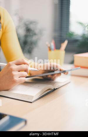 Nahaufnahme einer jungen Frau, die ihr Smartphone benutzt, während sie über offenes Buch am Schreibtisch studiert und den Stift hält, um Notizen zu machen. Mobiles Lernen, Online-Forschung, digital Stockfoto