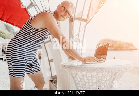 Freelancer Beach Laptop Mann in einem gestreiften Vintage Badeanzug, der draußen auf einem Tisch in der Nähe des Ozeans während der Sommerferien arbeitet Stockfoto