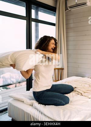 Frau spielt mit Kissen auf dem Bett in einem modernen Zimmer mit großen Fenstern an sonnigen Tagen Stockfoto