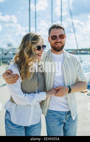 Ein Paar steht in der Nähe des Yachthafens, lächelt und genießt ihre Zeit. Sie tragen legere Kleidung und Sonnenbrille, während die Sonne hell scheint und Boote s Stockfoto