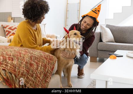 Paare, die auf dem Sofa sitzen und goldene Retriever streicheln, mit roten Hörnern, Mann mit Kürbiskegelhut Stockfoto