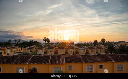 Sonnenuntergang erfüllt den Himmel über gemütlichen mediterranen Stadthäusern und Palmen. Die Sonne geht am Horizont nieder und wirft warmes Licht über die Dächer als Wolken dri Stockfoto