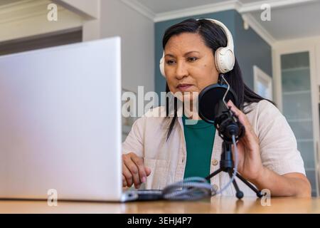 Reife Frau mit weißen Kopfhörern, die während der Aufnahme auf einem Laptop zu Hause in das Mikrofon sprechen Stockfoto