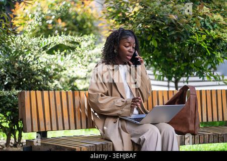 Beschäftigte afroamerikanische selbstständige Geschäftsfrau, die auf einem Laptop Multitasking und mit dem Smartphone spricht. Stockfoto