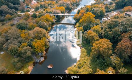 Aus der Vogelperspektive werden die lebhaften Farben des Herbstblatts um einen ruhigen Fluss und ein kleines Dorf, das von einer Fußgängerbrücke überquert wird, erfasst Stockfoto