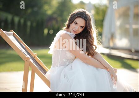 Wunderschöne Braut in einem weißen Kleid, die sich draußen auf einem Liegestuhl entspannt Stockfoto