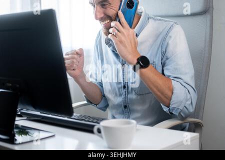 Glücklicher Mann, der am Schreibtisch seines Heimbüros vor einem Computer mit einem Smartphone spricht. Fernarbeit, Geschäftserfolg und positive Kommunikation Stockfoto