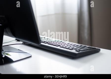 Minimaler Arbeitsplatz mit Computermonitor und Tastatur auf einem sauberen, weißen Schreibtisch, der von weichem Tageslicht beleuchtet wird. Modernes Büro und Remote-Arbeit Co Stockfoto