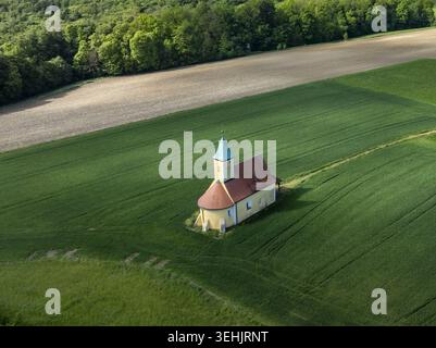 Aus der Vogelperspektive auf eine kleine historische Kapelle mit rotem Dach und grünem Turm, die inmitten üppiger grüner Felder in der Nähe eines walddeutschen Waldes steht Stockfoto