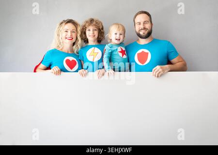 Familie von Superhelden, die zu Hause spielen. Vater, Mutter, Tochter und Sohn halten ein Banner frei. Eltern mit Kindern geben vor, Superhelden zu sein Stockfoto