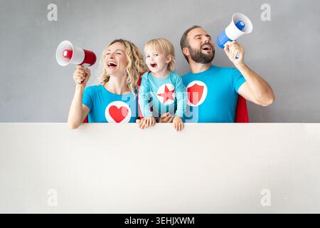 Familie von Superhelden, die mit Megaphon sprechen. Vater, Mutter und Kind halten Banner leer. Eltern mit Kind geben vor, Superhelden zu sein Stockfoto