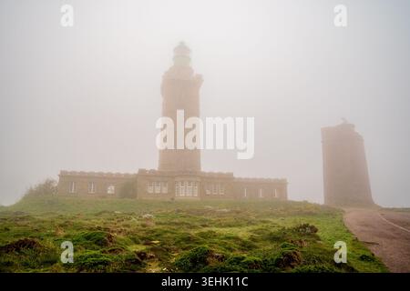 Ein flacher Blick auf den majestätischen Leuchtturm Cap Fréhel und seinen historischen Turm, umgeben von dickem Nebel in der Bretagne. Der grasbewachsene Boden und der Nebel kreieren Stockfoto