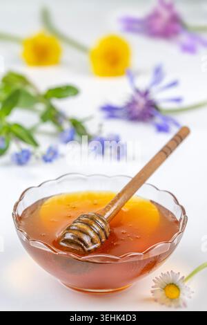 Eine Glasschale enthält eine viskose bernsteinfarbene Flüssigkeit und einen hölzernen Honiglöffel. Im Hintergrund und im Vordergrund befinden sich verschiedene Blüten. Stockfoto