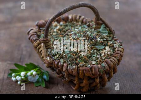 Ein gewebter Korb aus getrockneten Blättern und Blumen sitzt neben einem Zweig Maiglöckchen auf einer hölzernen Oberfläche. Stockfoto