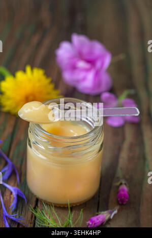 Ein Glas enthält Honig. Ein transparenter Kunststoffspachtel hält eine Portion Honig. Blumen sind im Hintergrund sichtbar. Stockfoto