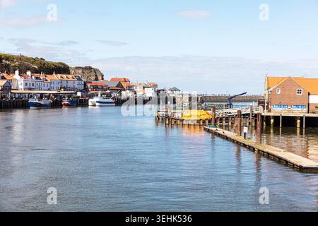 hafenszene in Whitby, North Yorkshire, zeigt Boote, die neben hölzernen Piers liegen, und ruhiges reflektierendes Wasser. Traditionelle Häuser und Geschäfte Stockfoto