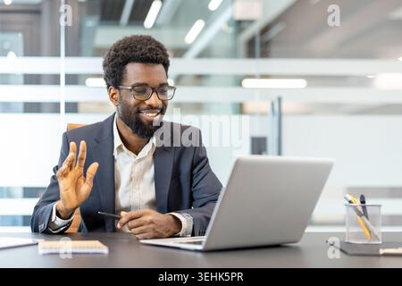 Professioneller Mann mit Brille lächelt und winkt während eines Videoanrufs auf einem Laptop, der die Fernkommunikation von einem modernen Büroarbeitsplatz aus wahrnimmt Stockfoto