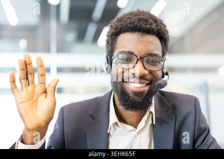 Professioneller afroamerikanischer Callcenter-Agent mit Headset und Brille, lächelnd und mit der Hand winkend, bietet Remote-Unterstützung und Unterstützung aus einer Büroumgebung Stockfoto