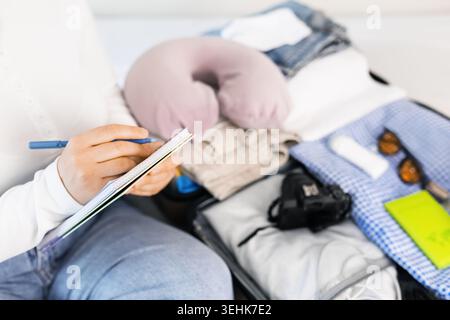 Nahaufnahme einer Person, die eine Packliste in ein Notizbuch neben einem offenen Koffer schreibt. Vorbereitung auf Sommerreisen, Urlaub und Tourismus mit Gepäck Stockfoto