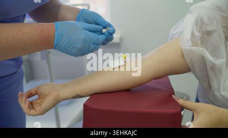 Nahaufnahme von medizinischem Fachpersonal in sterilen Handschuhen, das IV-Kanüle in die Vene am Arm der Frau einsetzt. Der Patient legt den Arm für den medizinischen Eingriff auf das rote Pad. Leuchtend Stockfoto