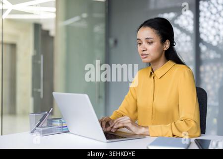 Junge Geschäftsfrau in eleganter Freizeitkleidung, die auf einem modernen Laptop an einem eleganten Schreibtisch schreibt und sich auf Arbeit und Produktivität in einem modernen Firmenumfeld konzentriert Stockfoto