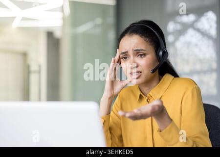 Junge schwarze Callcenter-Mitarbeiterin, die frustriert ist und Burnout erlebt, während sie an ihrem Schreibtisch arbeitet und einen Kunden per Headset und Laptop berät Stockfoto