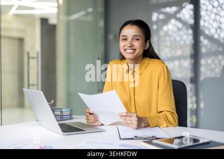 Junge professionelle Frau, die an einem Schreibtisch in einem modernen Büro sitzt, Dokumente in der Hand hält und die Kamera lächelt, um Vertrauen und Erfolg in einem Unternehmensumfeld zu vermitteln Stockfoto