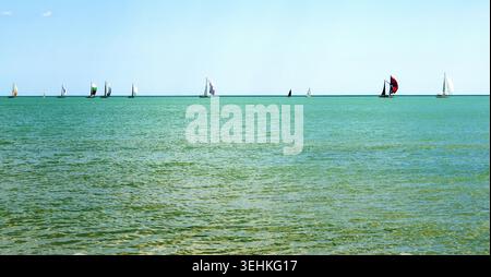 Mehrere Segelboote fahren über türkisfarbenes Wasser in Richtung des fernen Horizonts unter einem klaren blauen Himmel und fangen den Nervenkitzel des Segelns im Sommer ein Stockfoto