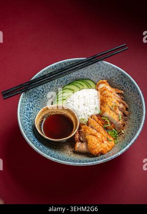 Ein Schuss knusprig gerösteter Enten- und Schweinebauch, serviert mit weißem Reis, Gurkenscheiben und Dippsoße in einer blauen Schüssel mit schwarzen Essstäbchen. Stockfoto