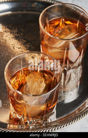 Nahaufnahme von Whisky Bourbon mit großen Eiswürfeln in zwei Kristallgläsern auf einem silbernen Serviertablett, warme Bernsteintöne und Reflexionen für Bar oder C Stockfoto