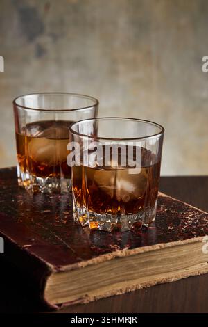 Zwei Gläser Bourbon Whisky mit Eiswürfeln ruhen auf einem alten ledergebundenen Buch auf einem Holztisch, mit rustikalem Hintergrund und W Stockfoto