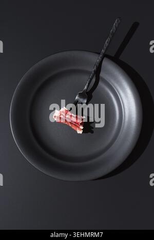 Minimalistisches Stillleben über dem Kopf eines schwarzen Tellers und einer Gabel mit dünnen Scheiben spanischen Schinken, isoliert auf einem dunklen Hintergrund mit dramatischen s Stockfoto