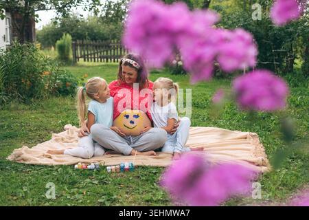Lächelnde schwangere Frau sitzt auf einer Decke in einem grünen Garten und umarmt ihre beiden Töchter. Niedliche Bauchmalerei und warme Familienbindung fangen fröhliche Kameraden ein Stockfoto