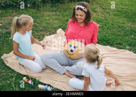Lächelnde schwangere Frau sitzt auf einer Decke in einem Park, während zwei Kinder auf ihren Babypuffer zeichnen, um Schwangerschaft, Familienbindung und fröhliche Outdoor-Aktivitäten zu feiern Stockfoto