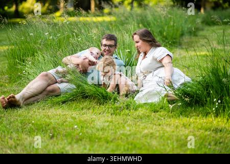 Lächelnde Eltern entspannen sich auf grünem Sommergras mit ihrem Baby und ihrer Tochter, teilen Liebe, Lachen und unbeschwerte Familienzeit im Freien in A Stockfoto