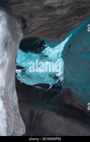 Abstrakter Blick in eine isländische Gletschereishöhle, mit blauen durchscheinenden Wänden, dunklen vulkanischen Schichten und natürlichem Licht, das durch gefrorene Korridore scheint Stockfoto