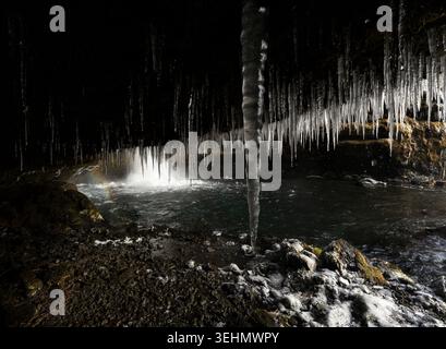 Moody isländische Höhlenszene mit einem winterlichen Wasserfall und Nebel, langen Eiszapfen, die über einem dunklen Bach von der Decke hängen, felsigem Boden und dramatischem tief Stockfoto