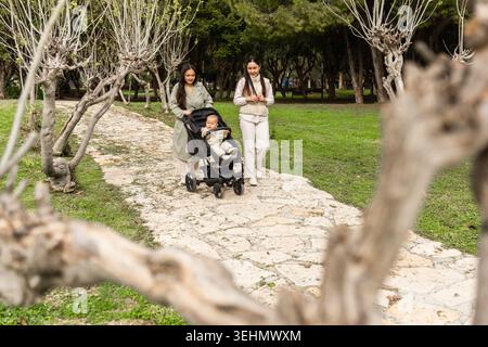 Eine Familie macht einen gemütlichen Spaziergang im Park. Zwei Frauen gehen einen Weg entlang, eine schiebt ein Kind in einen Kinderwagen. Die grüne Umgebung schafft eine ruhige Atmosphäre Stockfoto