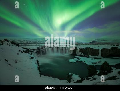 Grüne Nordlichter leuchten über einem gefrorenen Wasserfall Canyon und einem schneebedeckten Fluss in Island, gefangen in einer klaren Winternacht mit dramatischem Himmel und schroffen l Stockfoto