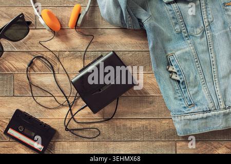 Flacher Retro-walkman ® Kassettenspieler mit orangefarbenen Kopfhörern, Jeansjacke und Sonnenbrille auf Holzbrettern, die an analoge Musik der alten Schule erinnern Stockfoto