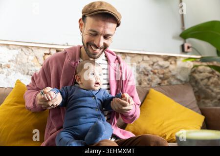 Der lächelnde Vater hält ein neugeborenes Baby auf einem gemütlichen Sofa zu Hause und teilt dabei süße Momente mit der Familie. Warme Lifestyle-Szene von neuen Eltern, Liebe, Pflege, Stockfoto