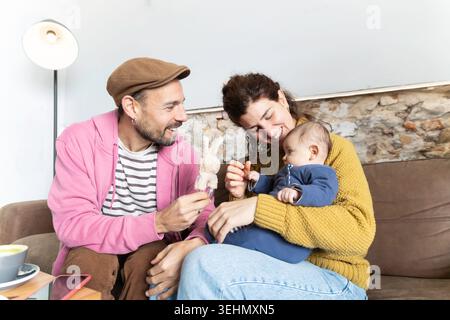 Glückliche Familie, die zu Hause süße Momente teilt, lächelnde neue Mutter hält ein neugeborenes Baby, während der Vater sanft spielt, Liebe, Bindung und frühe Eltern zeigt Stockfoto