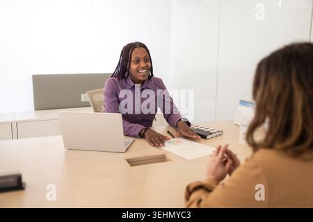 Ein lächelndes Coworking-Teammitglied führt eine Diskussion über einen Schreibtisch, verwendet einen Laptop und Papierkram in einem modernen Büro, schlägt Zusammenarbeit vor, plant eine Stockfoto