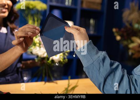 Eine Nahaufnahme eines Floristen und Kunden, die einen blauen Umschlag an einem Blumenladen austauschen. Auf der Rückseite sind helle, farbenfrohe Blumenarrangements zu sehen Stockfoto