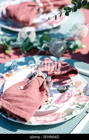 Dekoration des Frühlingstisches mit Blumentellern, rosa Leinenservietten und rustikalen Serviettenringen. Sonnendurchflutete Speiseszene im Freien mit Pastelltönen, Besteck, Stockfoto